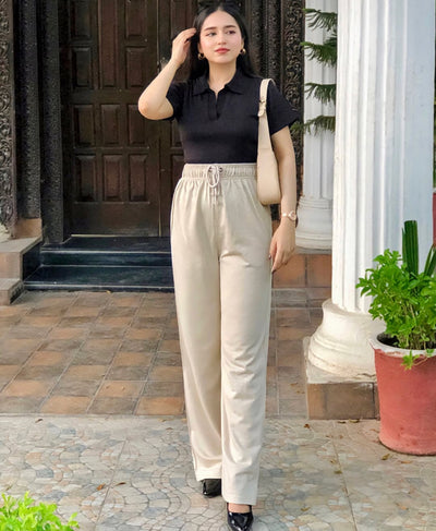 Woman in black top and beige pants standing outdoors with a plant and building entrance in the background